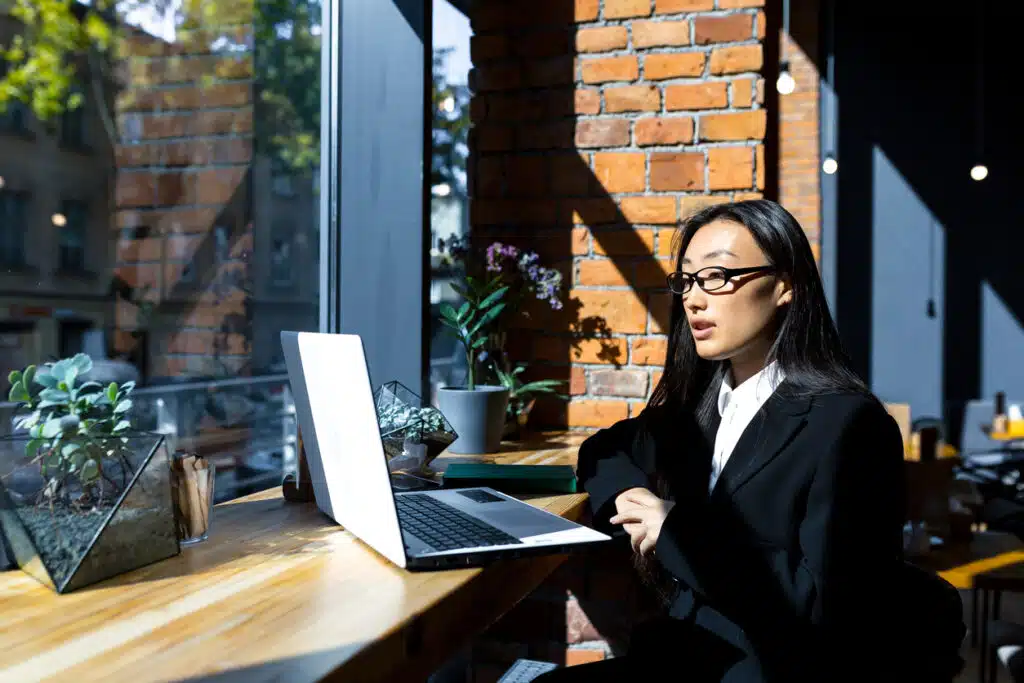 Business woman freelancer working in a cafe on a laptop remotely, pensive and focused Asian woman talking on a video call with colleagues, looking at laptop webcam virtual interview tips